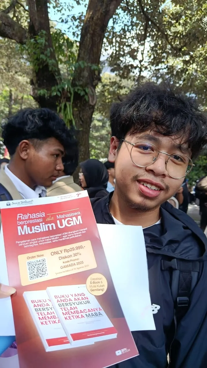 Daffa holds up the guidebook's printed flyer, with its order QR code, beside a fellow student at a crowded outdoor campus event.