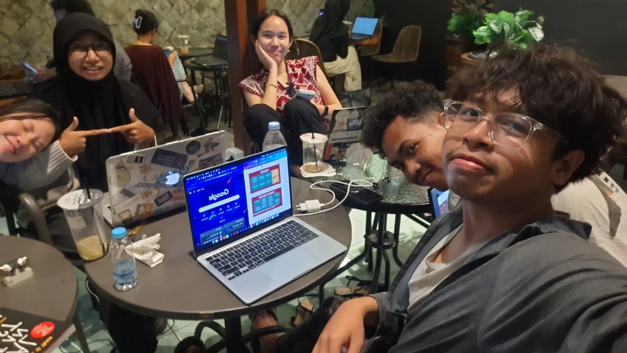 Behind the scenes, six group members smile for a selfie around café tables, laptops open in front of them.